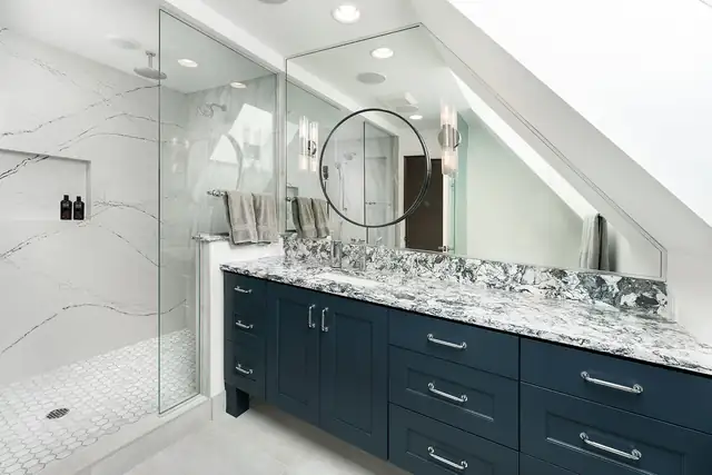 Primary bathroom with a navy blue vanity, marble-look counter, round mirror and a glass-enclosed shower under a sloped ceiling